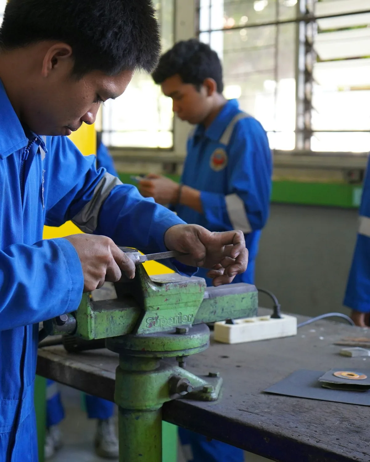 Trainees in overalls working at a bench vice in a training workshop